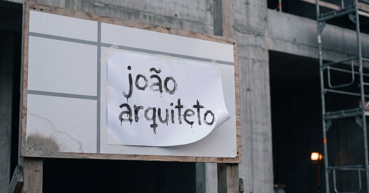 Placa de obra com os campos vazios e uma folha de papel presa torta com fita, com o nome de um escritório de arquitetura pintado à mão com pincel, na frente de uma estrutura de concreto inacabada