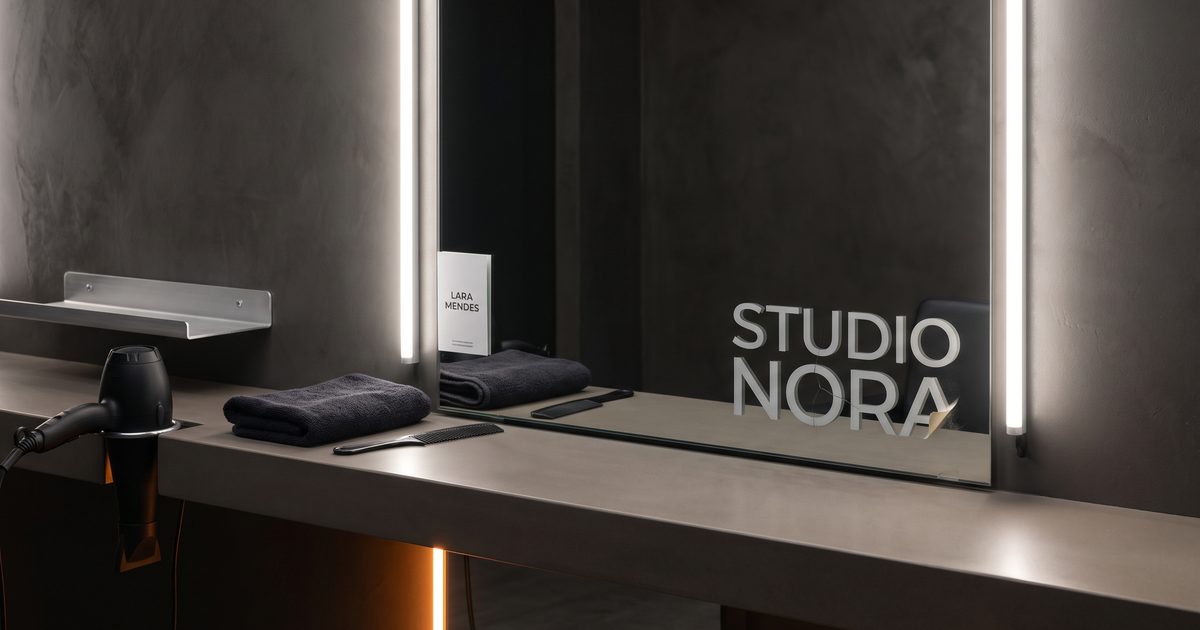 Espelho sem moldura numa parede escura de salão, com um adesivo de vinil recortado escrito STUDIO NORA no canto de baixo, com a quina descolada, e um cartão de visita novo escrito LARA MENDES encaixado em pé na borda do espelho